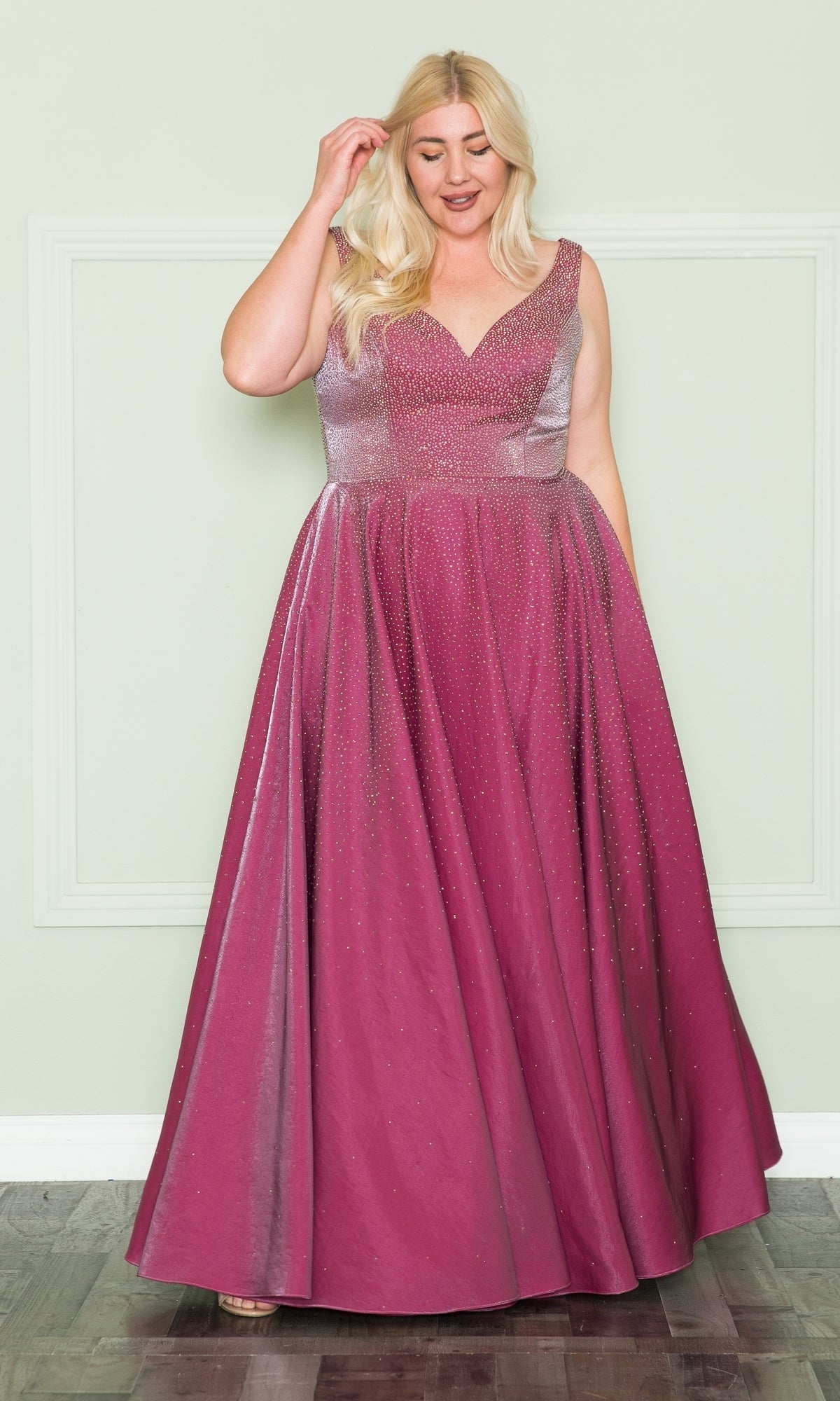 A woman wears the Embellished Plus-Size Long V-Neck Prom Dress W1110, a sleeveless, pinkish-purple gown, standing on a wooden floor before a pale green wall.